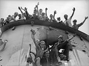 Close up of a ship's funnel, which has a large hole in the side. Sailors are smiling and waving at the photographer from the top of the funnel and inside the hole.