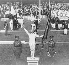 Photographie montrant Victor Boin dans le stade olympique levant le bras droit et tenant un drapeau dans la main gauche, entouré de deux soldats, devant les délégations.