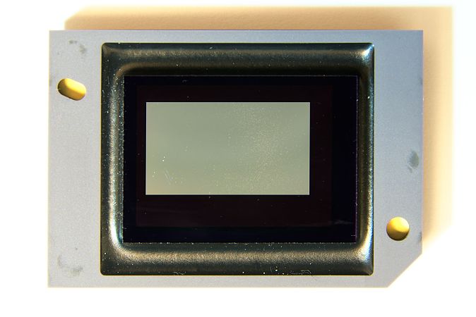 English: DMD chip from a videoprojector. View of the "white dots" among the micromirrors, on the right side. (About 3.2 × 2.2 cm (1.2 × 0.8 in)) Français : Puce DMD issue d'un vidéoprojecteur. Vue des « points blancs » dans les micro-miroirs, sur le coté droit. (Environ 3.2 × 2.2 cm (1.2 × 0.8 in))