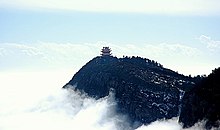Farbfotografie eines teilweise schneebedeckten Bergfelsens mit einem chinesischen Tempel an der Spitze. Unten links steigen Wolkenschwaden empor.