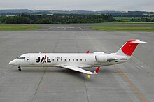 A Bombardier CRJ-200 aircraft taxiing on the tarmac, with a view of the runway and some bushes in the background