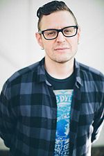 Kevin Kadish in a black t-shirt and unbuttoned checkered shirt wearing glasses