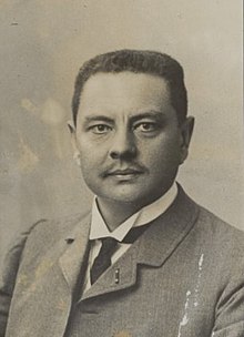 Portrait d'un homme moustachu portant un costume et une cravate.