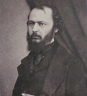 Fotografia em preto e branco de um homem barbudo posando em pé com a mão esquerda sobre o estômago.