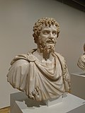 Bust of a man with curly hair and beard. He glances to his left. He wears a tunic and fringed cloak, fastened at his left shoulder with a circular brooch.