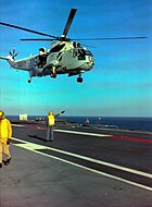 A helicopter hovering over the flight deck of a ship. Two men in fluorescent yellow vests are visible; one is using hand signals to direct the helicopter, the other is walking out of frame.