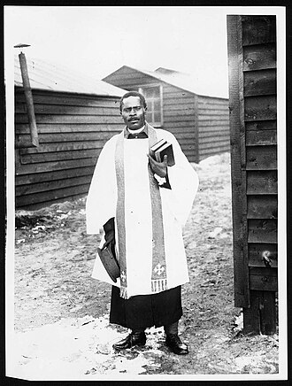 African priest, Western Front. He wears what seem to be Church of England vestments over army uniform. He is standing in a camp of wooden huts, probably one of the camps of the South African Native Labour Corps (National Library of Scotland 4687906729) African priest at Western Front during First World War.jpg