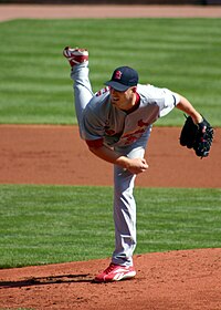 A man in a grey baseball uniform stands bent over with his right leg in the air having just pitched the ball to home plate.