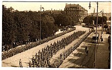 Schwarzweißfotografie in der Obersicht von einer Armee, die auf einer Straße von links unten nach rechts oben marschiert. An beiden Seiten ist ein Spalier mit Soldaten, die die Bevölkerung abgrenzt. Im linken Hintergrund sind Bäume und Menschen. An der rechten Seite sind Straßenbahn und Menschen. Im rechten oberen Bereich sind große Gebäude.