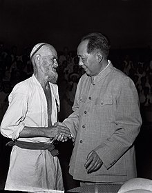 An elderly bearded Uyghur man shakes hands with a middle-aged Chinese man.