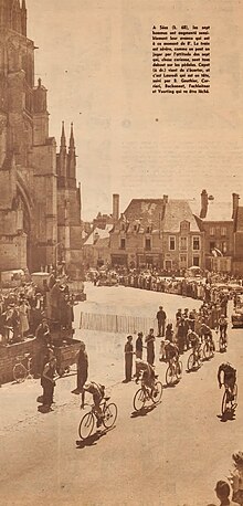 Photo en noir et blanc d'un groupe de cyclistes du Tour de France passant dans un village avec une église dans le fond de l'image.