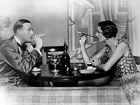 young white man, clean shaven, facing young white woman with bob cut hair across a small round table set with wine glasses and demitasse cups; both are smoking using cigarette holders