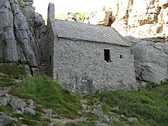 Photo d'un petit bâtiment presque cubique au pied d'une falaise, avec des murs épais et une toute petite fenêtre