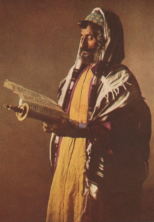 A Yemenite Jew in traditional vestments under the tallit gadol, reading from a Torah scroll