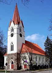Seitliche Farbfotografie einer Kirche mit weißem Turm und roter Spitze. Die Spitze hat neben dem großen Turm vier kleine Ecktürme. Der untere Bereich besteht aus dunkelbrauner Steinmauer und das weiße Kirchenschiff hat ein rotes Dach.