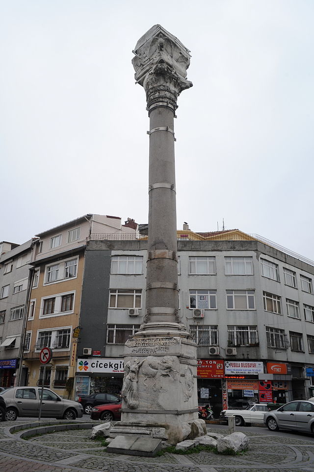 Color photograph of an ancient stone column set in front of a modern building and parked cars. The bottom and top of the column are engraved, and several metal bands placed at regular intervals encircle the central section of the column.