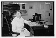 A middle-aged white woman in a nurse's uniform, seated at a desk