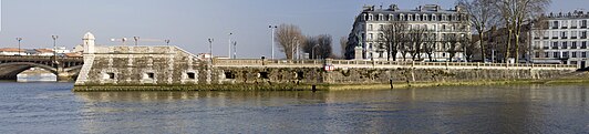 Vista de un muelle fortificado, adornado con una torre de vigilancia, a modo de prolongación de un puente sobre un arroyo.  A la derecha de la vista se encuentran dos imponentes edificios.