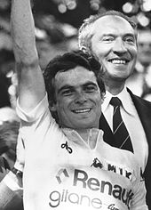 Black-and-white photo of Hinault in a cycling jersey, smiling and lifting his right arm in the air