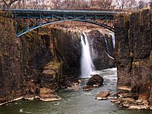 Les gorges d'une rivière, qu'enjambe un pont métallique. Derrière le pont, une chute d'eau.