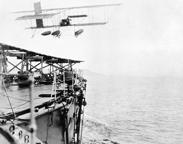 The first take-off by an aeroplane from a moving ship, 2 May 1912. Hibernia's bow and flying-off ramp at bottom left.