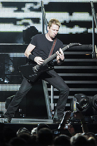 A blonde man wearing a dark T-shirt and dark jeans playing a guitar on a stage.