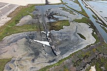 The wreckage of a large jet aircraft lying in a marsh, facing the bottom of the frame. The fuselage is broken in front of the tail, and the fuselage and left wing have been gutted by fire damage. Only one engine is still attached; it lies under and in front of the aircraft's right wing. Several large furrows extend backwards from the aircraft towards the top of the frame. Paved surfaces are visible at extreme top center and top left. Many scattered pieces of wreckage lie behind the aircraft in the furrowed areas.