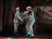 Deux hommes en costume-cravate à carreaux vert et blanc et petit chapeau, sur scène, qui jouent respectivement du piano et du fiddle (petit violon) avec un grand sourire forcé.