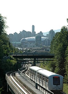 A two car train follows rail tracks under and bridge. In the background can be seen a domed sports stadium and high-rise buildings.