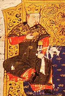 A man sits on a couch in front of a golden screen, wearing brown and red robes and a gold crown.