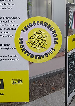 Ein rundes Schild an einer Infotafel weist darauf hin, dass die Ausstellung rassistische, antisemitische und gewaltvolle Inhalte zeigt.