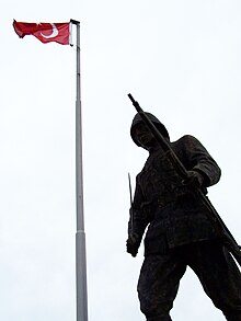 Photographie en contre-plongée d'une statue en bronze représentant un soldat tenant un fusil et un sabre. Un drapeau turc au sommet d'un mat est visible à l'arrière-plan.