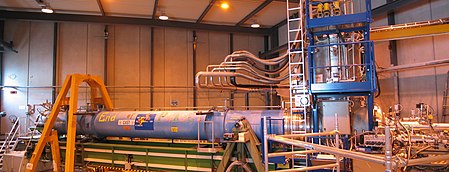 A warehouse interior containing a long blue cylinder surrounded by scaffolding and plumbing.