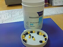 White bottle on blue pad atop a desk. The bottle cap is off, and is upside down on the pad in front of the bottle. In the cap are a dozen black-and-yellow capsules.