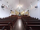 Fotografía en color del interior de una iglesia sin pasillo ni crucero, totalmente pintado en blanco, con bancos de madera.