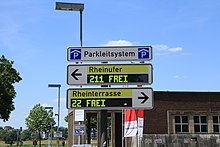 View of three elongated white signs.  At the top with black writing “Parkleitsystem”, on both sides a blue rectangle with a white P under a white roof.  The sign underneath points with a black arrow to the left, next to it stands on a yellow background "Rheinufer".  A rectangular black display is arranged under this black lettering.  It shows “211 FREE” with a blocky, light yellow font.  The sign below has the same structure, but points to the right.  The writing reads "Rheinterrasse" at the top, "22 FREI" at the bottom.  Behind the signs you can see the sky, some trees and buildings.