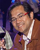 A man with dark hair and glasses, holding a glass of wine and smiling at the camera.