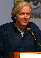 A man in a blue jacket, with a gray shirt underneath, in front of a microphone. The eye logo for the San Diego Comic-Con is seen in the background.