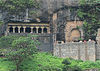 A rock face with a six-pillared cave front (left) and a carved cave entrance (right). Greeny patch at the bottom.