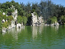 Rocky, bushy bluffs at the rear of some green water. There is a small structure with a Chinese roof at the base of one, in the center