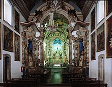 Photographie en couleurs. Intérieur de la nef : les murs sont blancs et affublés de nombreux tableaux ; les angles sont forces de reliefs en couleurs, très baroques, tandis que l'autel au fond semble illuminé d'une lueur verte.