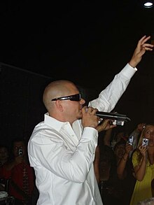 Side view of a man with a shaved head, wearing sunglasses and a white dress shirt at a high school prom. He holds a microphone with his right hand to his mouth and his left hand is held up. Behind him, several students are seen taking photographs with digital cameras and cell phones.