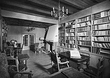 A black and white image of a room with a wood panelled ceiling, with a large fireplace and bookshelves on two sides of the room. At the far end of the room is a glass fronted double door leading away. There are a number of small chairs and tables around the room.