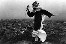 Photographie en noir et blanc, montrant une Sœur de la Perpétuelle Indulgence en costume, une fleur à la main, sur les hauteurs d'une colline.