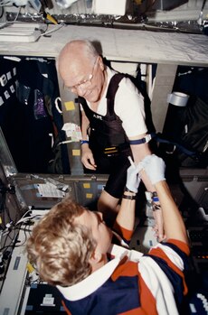 Glenn, wearing his glasses and black coveralls over a white T-shirt, has the inside of his elbow taped by a crew member wearing an orange and blue polo