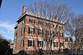 A three story brick house with white trim and black shutters. A wrought iron fence surrounds the front yard, and there is a low railing surrounding the roof.