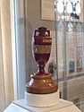 A terracotta urn with two brass plaques on it sits on a wooden base. The urn is on display, with the glass surround and other exhibits visible in the background