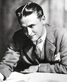 Una fotografía de F. Scott Fitzgerald por Nickolas Muray.  Fitzgerald está inclinado sobre un escritorio y examina un fajo de papeles.  Lleva un traje ligero y una corbata de lunares.  Un pañuelo blanco está en el bolsillo del pecho.