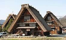 Színes fotó két fából készült hegyi faház típusú házból, nádfedeles tetővel, kék ég háttérrel.