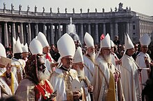 Bishops vested in white are standing in the sunshine in St. Peter's Square. Most wear white mitres on their heads, except for an Eastern Catholic bishop who wears a distinctive embroidered velvet hat.